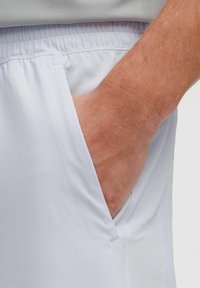 Main droite insérée dans la poche avant d'un short à taille élastique gris clair sur un fond neutre.