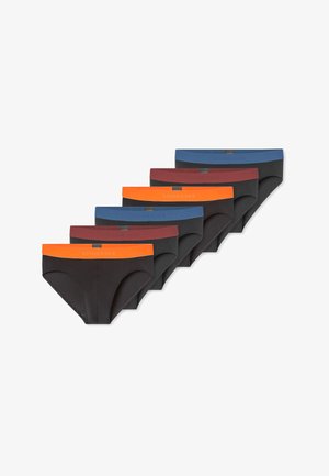 Junger Mann mit kurzen dunklen Haaren, der schwarze Slips mit einem orangefarbenen Taillenbund trägt, steht und blickt seitlich vor einem einfarbigen Hintergrund.