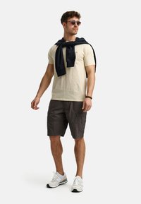 Beige bomulds t-shirt, grå cargo shorts, marineblå sweater draperet over skuldrene, grå og hvide sneakers, solbriller, afslappet look.