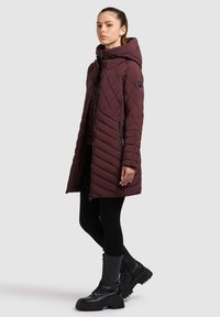 Geacă matlasată bordo cu glugă, prevăzută cu fermoar și buzunare laterale, purtată peste colanți negri și cizme negre masive.