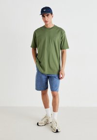 Grøn bomulds T-shirt, afslappet pasform, korte ærmer, diskret logo. Sammen med denim shorts og beige sneakers med hvide sokker. Blå kasket bæres.