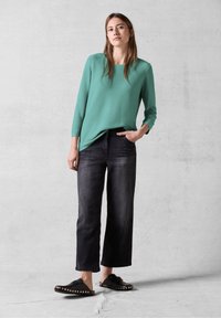 Türkiser Langarmshirt mit lockerer Passform, kombiniert mit dunkelblauen Schlagjeans und schwarzen Slippers mit goldenen Nieten.