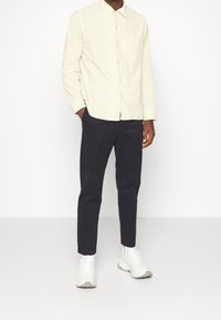 Jasnobeżowa koszula na guziki z kieszenią na piersi, zestawiona z ciemnogranatowymi spodniami i białymi butami sportowymi z szarymi akcentami.