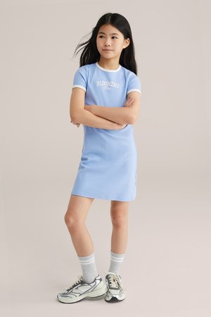 Dívka stojící se založenýma rukama, oblečená v světle modrých šatech s nápisem "BLOOMING", šedé ponožky a bílo-černé tenisky na jednobarevném pozadí.