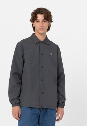 Veste légère grise avec un col, fermeture à boutons, poignets élastiques et poches latérales. Présente un petit logo sur la poitrine.