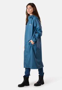 Długi niebieski wodoodporny płaszcz przeciwdeszczowy z zatrzaskami, bocznymi kieszeniami, regulowanym kapturem, w połączeniu z czarnymi gumowymi bootami i niebieskimi dżinsami.