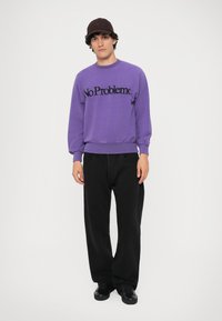 Lilla sweatshirt med "No Problemo" i sort, kombineret med baggy sorte bukser og sorte sko, båret med en mørk kasket, stående på en hvid baggrund.