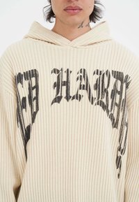 Cremefarbene, gerippte Kapuzenjacke mit auffälligem schwarzen Text, lockerem Schnitt und Kordelzugkapuze. Der Stoff ist weich und leicht dehnbar.