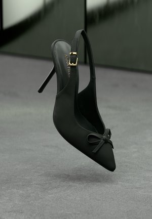 Black high-heeled slingback shoe with pointed toe, small bow on front, and gold buckle on ankle strap, displayed against a gray background.