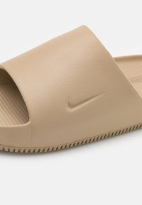 Beige slide sandal made of textured plastic with an open toe design. Features a small embossed Nike logo and a ridged sole for traction.