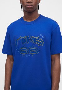Άτομο που φοράει μπλε βασιλικό t-shirt Nike SB με κείμενο με κίτρινο περίγραμμα και μικρά λευκά αστέρια στο μπροστινό μέρος.