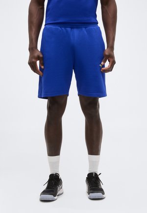 Άνδρας που φοράει μπλε αθλητικά σορτς, λευκές κάλτσες μέχρι τον αστράγαλο και μαύρα παπούτσια για τρέξιμο, στέκεται μπροστά σε απλό φωτεινό φόντο.