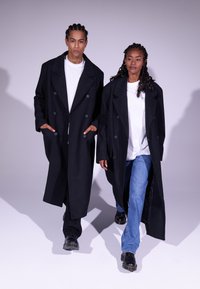 Manteaux noirs croisés à double boutonnage avec de gros boutons, portés par-dessus des t-shirts blancs et des jeans. Des bottes sombres complètent les tenues.