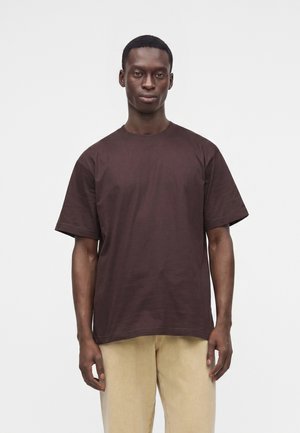 Uomo con capelli corti che indossa una maglietta semplice a maniche corte di colore marrone scuro e pantaloni beige chiaro, in piedi davanti a uno sfondo bianco.
