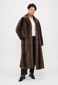 Hnedý kožuch z umelého vlneného materiálu so šálovým golierom, po kolená, s bočnými vreckami a hladkou textúrou. Nosí sa cez biele tričko a čierne nohavice.