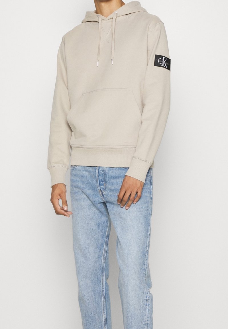Sudadera beige con un bolsillo frontal y capucha con cordón. Presenta un parche de logo negro en la manga izquierda. Combinada con jeans de color azul claro.