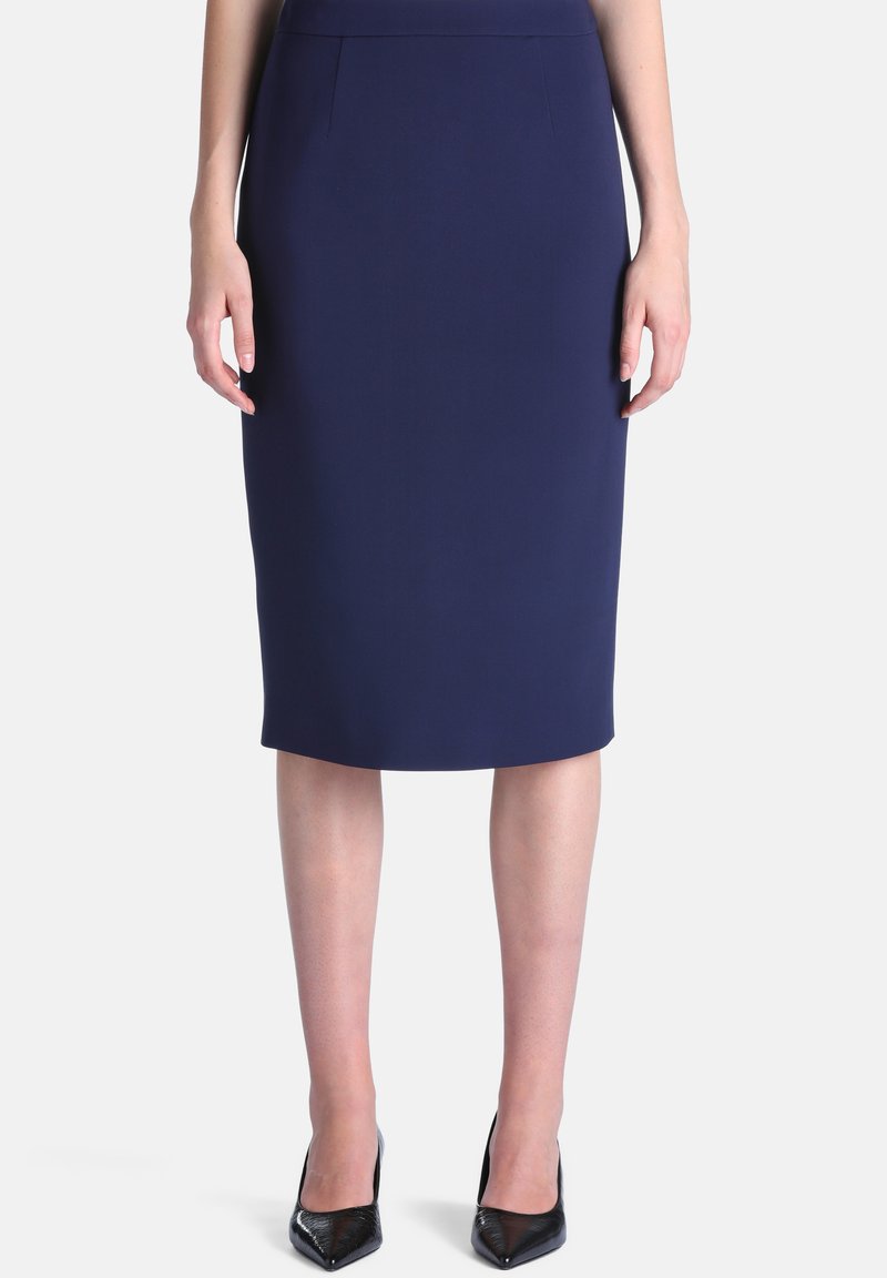 Femme portant une jupe crayon bleu marine longueur genou avec des talons hauts noirs à bout pointu, debout devant un fond blanc uni.
