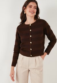 Cardigan rayé marron avec un tissu texturé, doté de six boutons ronds dorés. Manches longues et col rond. Associé à un pantalon beige.