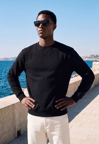 Homme portant un pull noir et des lunettes de soleil se tient les mains sur les hanches près de la promenade en bord de mer par une journée ensoleillée et claire.