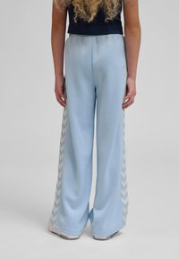 Person iført lyseblå bukser med vide ben og hvide og blå chevronstriber på siderne, stående mod en ensfarvet baggrund.