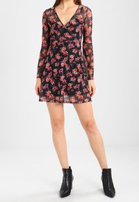 Schwarzes Blumenkleid mit langen transparenten Ärmeln, V-Ausschnitt und figurbetonendem Oberteil. Mit ausgestelltem Saum und kleinen rosa und roten Blumenmustern. Schwarze Knöchelstiefel.