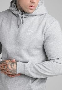 Sudadera de algodón gris con cordones y puntas plateadas. Presenta un logo bordado en el pecho y puños acanalados. Textura suave y corte regular.