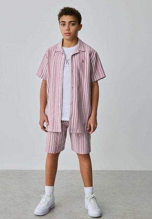 Băiat adolescent în picioare, purtând o cămașă cu mâneci scurte cu dungi verticale roz și albe și șorturi asortate, tricou alb cu imprimeu, șosete albe și adidași.