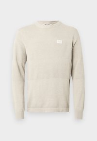 Lys beige strikkegenser med teksturerte mønstre, lange ermer og rund hals. Har en liten etikett på brystet.