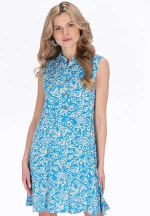 Femeie cu păr lung blond, purtând o rochie albastră fără mâneci, cu modele florale albe și nasturi pe partea din față, stând în fața unui fundal alb.