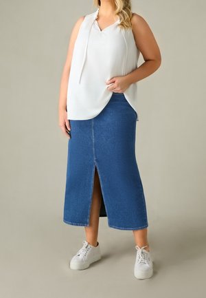 Kobieta ubrana w białą bluzkę bez rękawów, niebieską dżinsową spódnicę midi z rozporkiem z przodu oraz białe sneakersy na platformie, stoi na jednolitym tle.