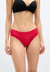 Γυναίκα που φοράει μαύρο σουτιέν και κόκκινα δαντελένια εσώρουχα, στέκεται μπροστά σε λευκό φόντο, με ορατό τον κορμό και τα πάνω μέρη των ποδιών.