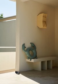 Tealfärgad uppblåsbar stol formad som en varelse med armar, vilande på en vit betongbänk mot en ljus vägg. Svarta skor på golvet.