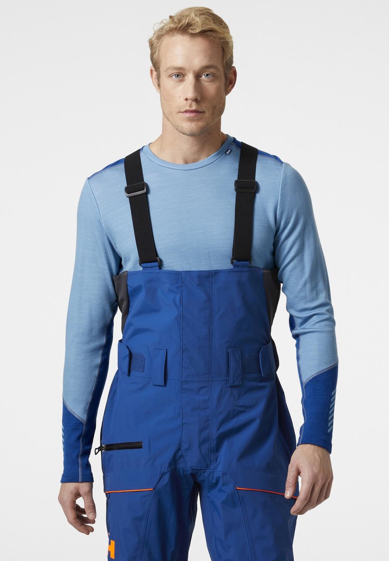 Modré nepromokavé kombinézy s nastavitelnými černými popruhy, texturovaný modrý vršek, zdobený zipy a jasně oranžovými detaily.