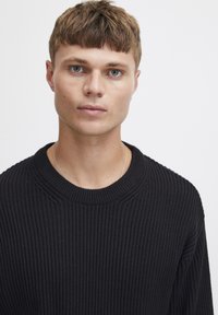 Pulover negru tricotat, cu textură în dungi și guler rotund. Stil ajustat, cu mâneci lungi, detalii minime și un material moale.