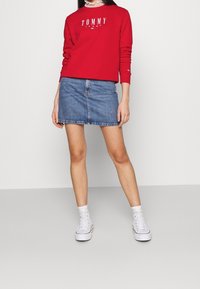 Červená mikina s logem "TOMMY JEANS", kombinovaná s krátkou modrou denimovou sukní. Bílé vysoké tenisky a bílé kotníkové ponožky dodávají outfitu šmrnc.