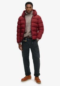 Doudoune rouge avec capuche, un pull à col roulé gris clair, un pantalon cargo foncé et des bottes à lacets marron. Tissu texturé et zips accentués.