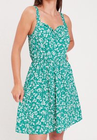 Robe fleurie verte avec motifs blancs, dotée d'un décolleté coeur, de bretelles ajustables et d'une taille élastique, fabriquée en tissu léger.