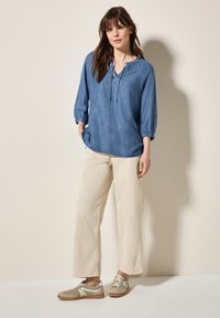 Camicia azzurro chiaro a maniche lunghe con scollo regolabile; abbinata a pantaloni larghi color crema e sneakers beige con dettagli bianchi.
