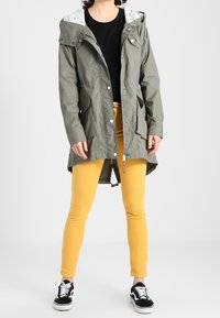 Oliwkowa kurtka z kapturem, zapinana na napy, z wzorzystą podszewką, zestawiona z żółtymi skinny jeansami i czarnymi sneakersami.