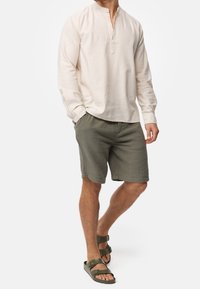 Mężczyzna w beżowej koszuli z długim rękawem typu henley, oliwkowych teksturowanych szortach oraz oliwkowych sandałach na podwójnych paskach, stojący z jedną ręką w kieszeni.