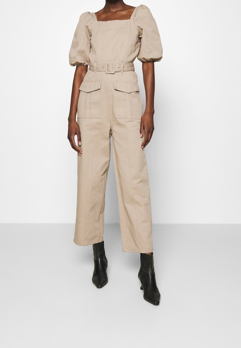Femme portant une combinaison beige ceinturée avec des manches bouffantes, un col carré, de grandes poches avant et des bottines noires.