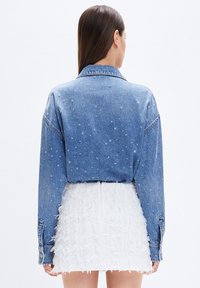 Giacca in denim con un lavaggio blu, caratterizzata da una vestibilità rilassata e da una leggera usura. Abbinata a una gonna bianca testurizzata.
