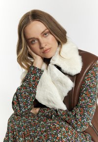 Bruine leren vest met een witte nepbont kraag, gecombineerd met een donkere jurk met bloemenpatroon met groene, rode en paarse bloemen.