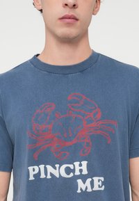 Μπλε T-shirt με κοντά μανίκια από βαμβάκι, με γραφικό κόκκινο κάβουρα και το κείμενο "PINCH ME" με έντονα λευκά γράμματα στο κάτω μέρος.