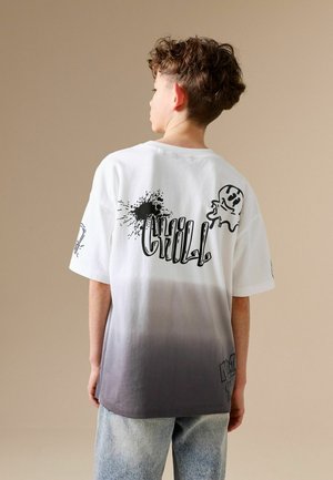 Chlapec obrácený zády, má na sobě tričko s bílým až šedým přechodem a černým grafitovým nápisem „CHILL“ a kreslenou postavičkou ducha na zádech.