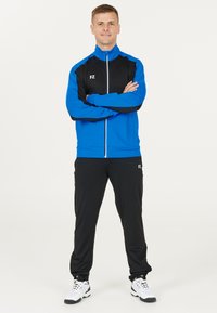 Mans sportkläder med en blå och svart zip-up jacka samt svarta byxor med vita accents. Materialet verkar slätt och andningsbart.