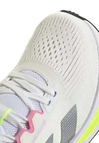 Zapatilla deportiva blanca con una parte superior de punto transpirable, paneles de malla, acentos en gris claro, detalles en rosa y amarillo, y una superposición texturizada en el costado.