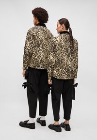 Beige és fekete leopárdmintás dzsekik, laza fekete nadrágokkal, melyeken masni díszítések vannak. Mindketten fekete cipőt viselnek, hátulról nézve.