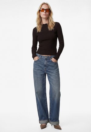 Pitkähiuksinen nainen, jolla on oranssinsävyiset aurinkolasit, musta pitkähiha paita, väljät siniset farkut käänteillä ja ruskeat teräväkärkiset korkokengät.