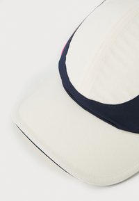 Boné com um tecido branco texturizado, faixa azul marinho e detalhe rosa de um lado. Viseira suave, design estruturado, adequado para uso casual.